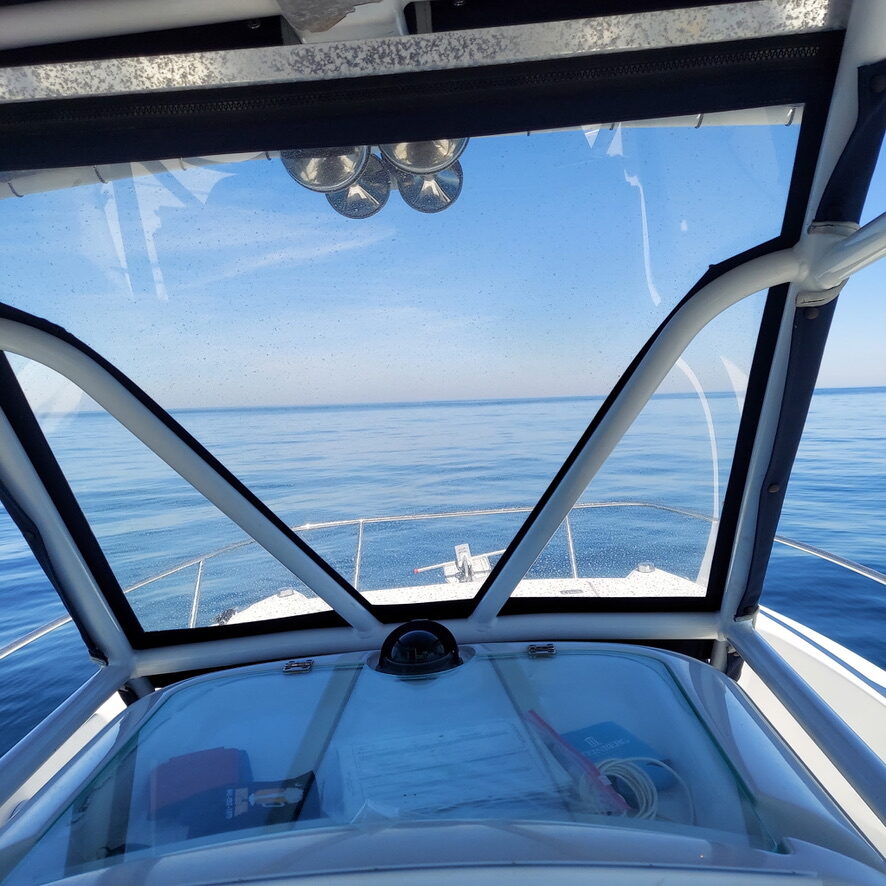 View from boat cabin through windshield toward calm open sea under clear blue sky.